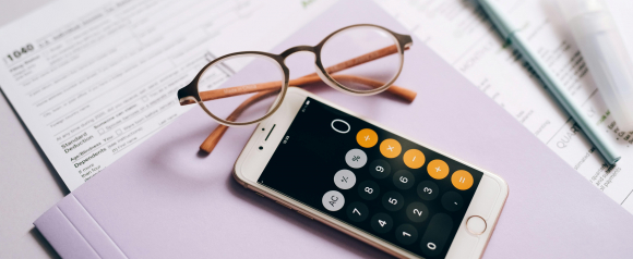 A smartphone with a calculator app open sits on top of a lavender folder, next to eyeglasses and partially visible IRS 1040 tax forms, representing financial planning and protection tax preparation.