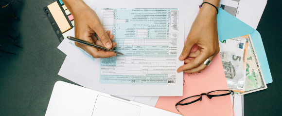 Close-up of hands filling out a U.S. 1040 individual income tax return form with a pen, surrounded by paperwork, colorful folders, euro and dollar bills, and eyeglasses on a dark desk. The scene reflects a financial planning or tax preparation setting, relevant to services offered by a tax group center.