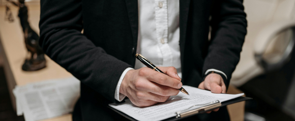 A person in a black blazer and white shirt signs a legal document on a clipboard, with a statue of Lady Justice blurred in the background. The professional setting suggests legal or tax-related services, fitting for Victory Tax Lawyers.