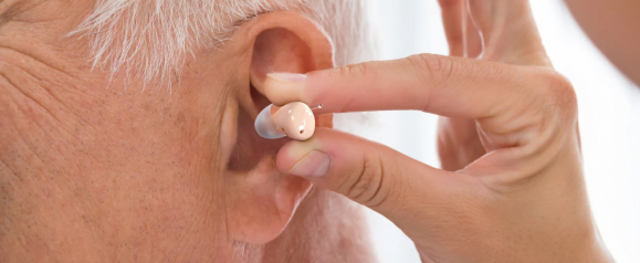 Close-up of a person inserting a flesh-toned Audien hearing aid into the ear of an older adult with gray hair. The in-ear device is small and discreet, emphasizing Audien’s nearly invisible hearing solution.