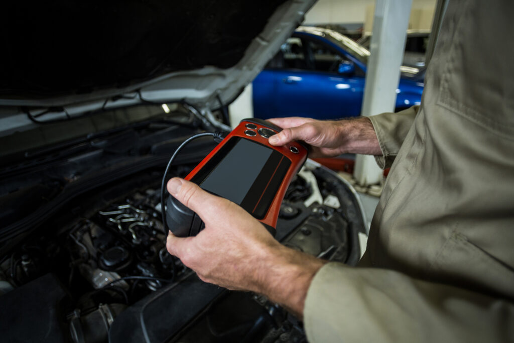 A mechanic holds a handheld diagnostic scanner connected to a car’s engine, running a system check through the vehicle’s OBD port. The open hood and visible engine components emphasize the diagnostic process in a garage setting.