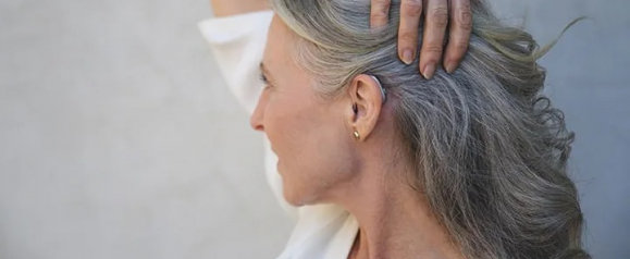 Close-up side profile of an older woman with long, wavy gray hair wearing a discreet Jabra Enhance hearing aid behind her ear. She lifts her arm and brushes back her hair, subtly revealing the device while wearing a white top and gold hoop earring.