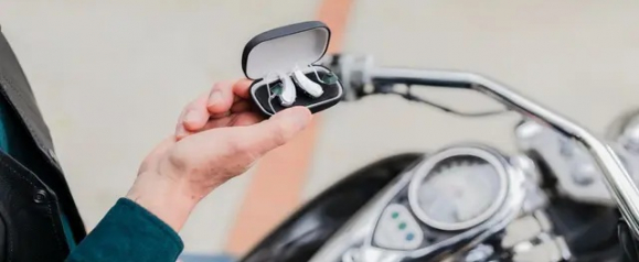 A person holding an open Lexie hearing aid case near a motorcycle handlebar, with two white in-ear hearing aids visible inside. The image highlights the portability and accessibility of Lexie hearing aids in an active lifestyle setting.