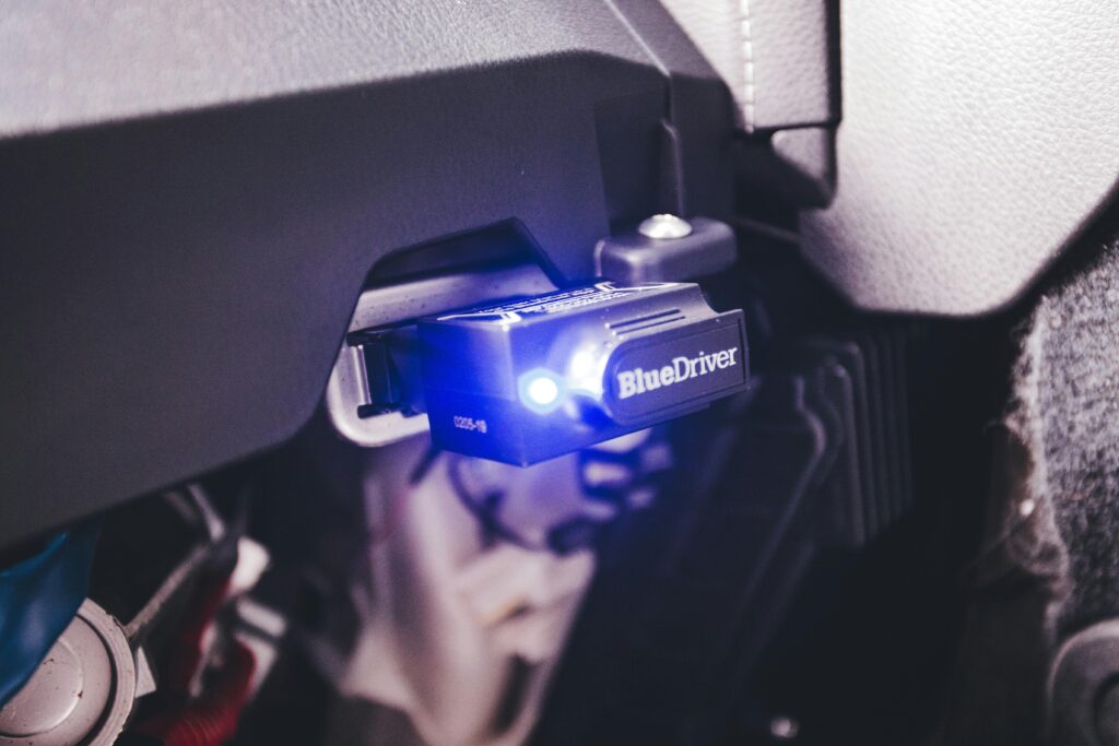 A BlueDriver diagnostic tool plugged into a vehicle’s OBD port beneath the dashboard, with a bright blue indicator light showing it’s powered on. The image highlights the device’s secure connection to the onboard diagnostics system.