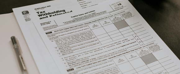 Close-up of IRS tax forms including Schedule D for Capital Gains and Losses and Publication 505 for Tax Withholding and Estimated Tax, with a pen and folder nearby. The image suggests the paperwork and detail involved in filing taxes, highlighting the potential need for expert help from services like Anthem Tax Services.