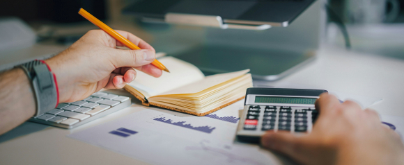 A person uses a calculator with one hand while writing in a notebook with the other, surrounded by a keyboard, documents, and financial charts. This workspace scene reflects the detailed tax and financial analysis offered by BC Tax.