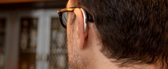 Rear view of a man wearing glasses and an over-the-ear hearing aid, with the device resting comfortably behind his ear. The image demonstrates the fit and design of AudioNova hearing aids in a casual, everyday setting.