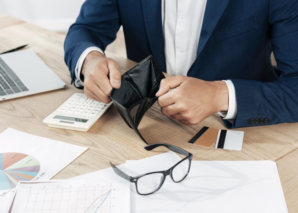 Man in a blue suit sitting at a desk, holding open an empty wallet with credit cards, financial charts, a calculator, and a laptop nearby. The image visually represents financial distress, supporting topics like bankruptcy and unresolved tax debt.
