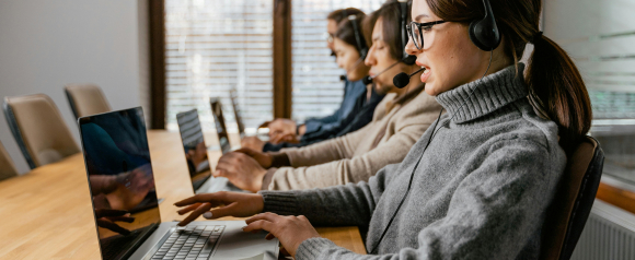 Four customer support agents wearing headsets sit in a row working on laptops in a modern office, with the focus on a woman in a gray turtleneck speaking into her mic. The image conveys a professional CRM team likely using Monday.com or similar software to manage client communications.