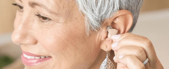 Close-up of a smiling older woman with short gray hair inserting a Nano hearing aid into her ear. She wears a silver earring and a ring, and her expression appears content and confident.