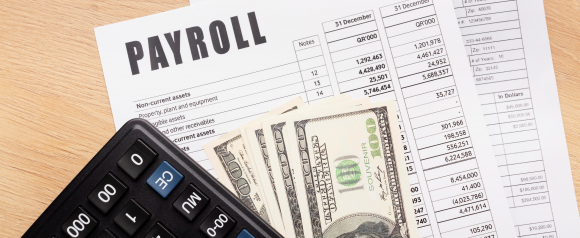 A payroll report labeled PAYROLL lies on a wooden desk alongside a calculator and a stack of US dollar bills partially covering detailed financial columns. The image visually represents payroll processing and employee compensation, supporting content about payroll software like Rippling used to manage pay calculations and records.