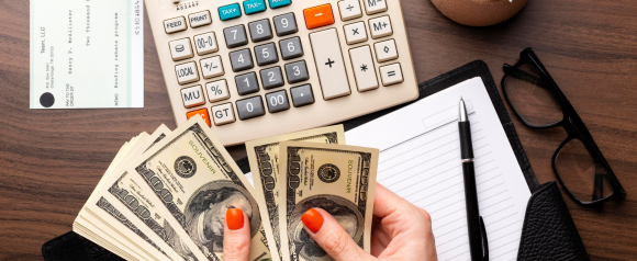 Hands with red nail polish count a stack of US dollar bills on a desk next to a calculator a business check a notebook and a pen suggesting payroll or financial processing. The image supports content about payroll software like Deel by visually representing employee payments budgeting and compensation management.