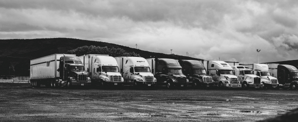 Black and white photo of a fleet of semi-trucks parked in a row on wet pavement under a cloudy sky, with visible company logos like TWT, Swift, and TransAm. The image highlights the scale and variety of commercial trucks, relevant to fleet tracking solutions like Spireon.