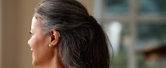 Side view of a woman with gray-streaked dark hair wearing a behind-the-ear Widex hearing aid, partially hidden by her hair. The image highlights the discreet and ergonomic design of the hearing aid in a natural indoor setting.
