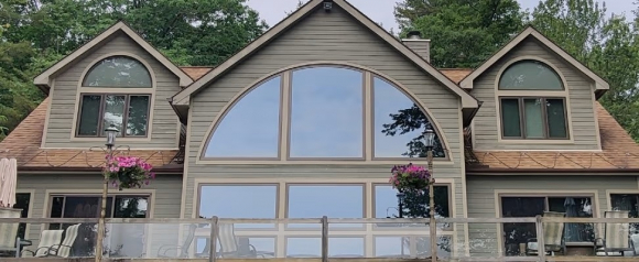 A lakeside home featuring large Renewal by Andersen windows, including a dramatic arched picture window framed by smaller rectangular panes. The reflection of trees and sky highlights the window clarity and energy-efficient design.