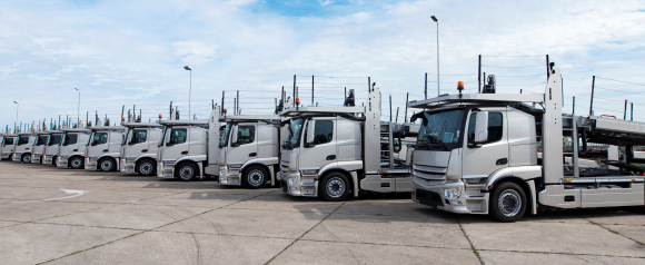 A row of identical silver car hauler trucks parked in a lot under a partly cloudy sky, lined up in perfect alignment. The image illustrates the scale and organization made possible with fleet tracking solutions like Geotab.