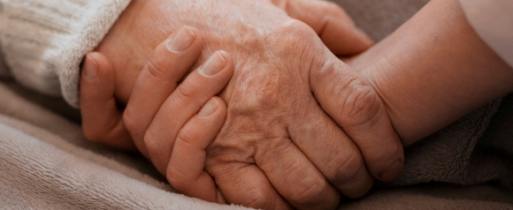 Close-up of a younger hand gently holding an older hand resting on a blanket, symbolizing comfort, caregiving, and support. The image reflects the emotional reassurance and safety that medical alert systems like Lifeline aim to provide for seniors.