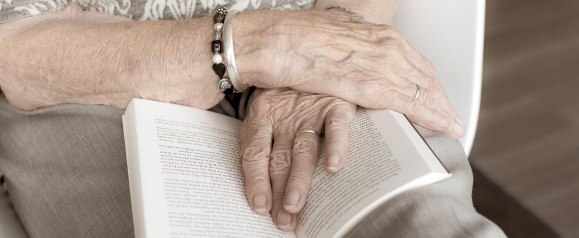 Close-up of an elderly person's hands resting on an open book, with one hand wearing a charm bracelet and the other a wedding band. The calm, reflective moment underscores the value of independence and how a Medical Alert system can help seniors maintain that freedom safely.