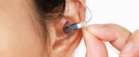 Close-up of a person inserting an AcoSound hearing aid into their ear, showing the slim, clear tubing and soft ear dome for a discreet and comfortable fit. The image highlights the precision and ease of use of the hearing device.