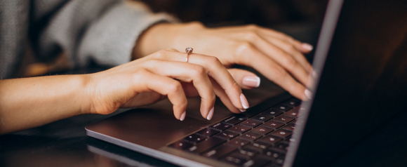 Close up of hands typing on a laptop keyboard with a silver engagement ring visible, representing everyday online activity like shopping banking or working remotely. The image highlights the importance of TotalAV antivirus software in protecting personal information and financial data while using a computer.