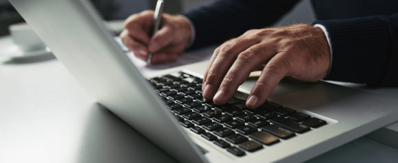 Close up of a person typing on a laptop while holding a pen and reviewing paperwork, representing everyday online work and financial tasks. The image highlights the importance of Avira antivirus software in protecting sensitive information and business data during daily computer use.