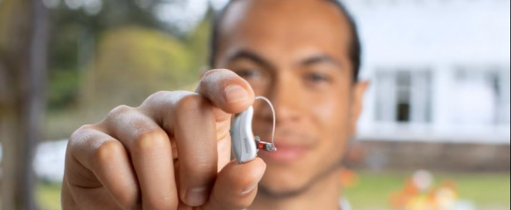 Man holding a small behind the ear hearing aid between his fingers with a home and garden blurred in the background. The compact device features a curved wire and discreet design, highlighting modern hearing technology. The image showcases Phonak hearing aids as a reliable and advanced solution for clearer everyday sound.