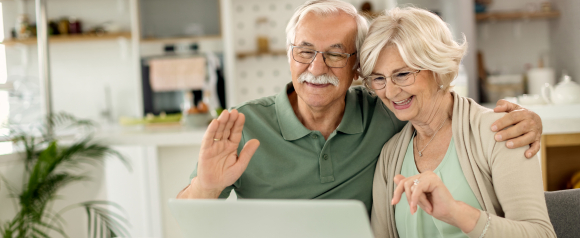 Happy senior couple using a laptop during breakfast, waving and smiling on a video call, showing how Telus LivingWell Companion and medical alerts support connected, independent living.