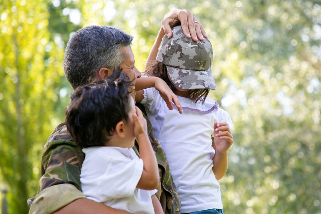 Military parent in camouflage holding two young children outdoors while one child adjusts a camouflage cap. The warm family moment reflects the everyday life and financial realities faced by many military families.