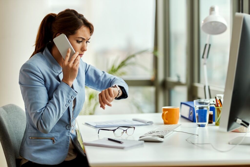 Businesswoman on a phone call checking her watch while working overtime at her desk