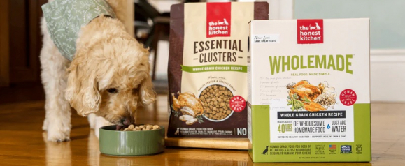 Dog eating from a bowl next to The Honest Kitchen Essential Clusters and Wholemade dog food packages