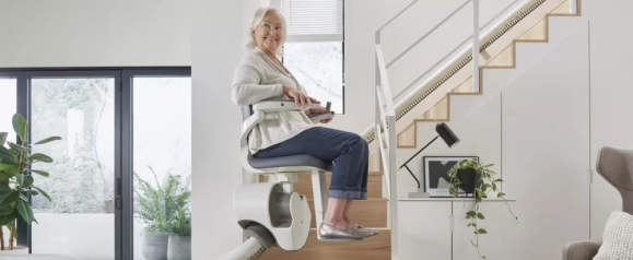 Smiling elderly woman riding a stairlift chair along a modern home staircase, holding the remote control