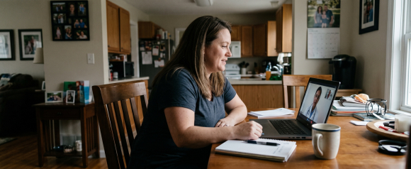Woman taking notes during a telehealth consultation with a doctor on her laptop at a home kitchen table