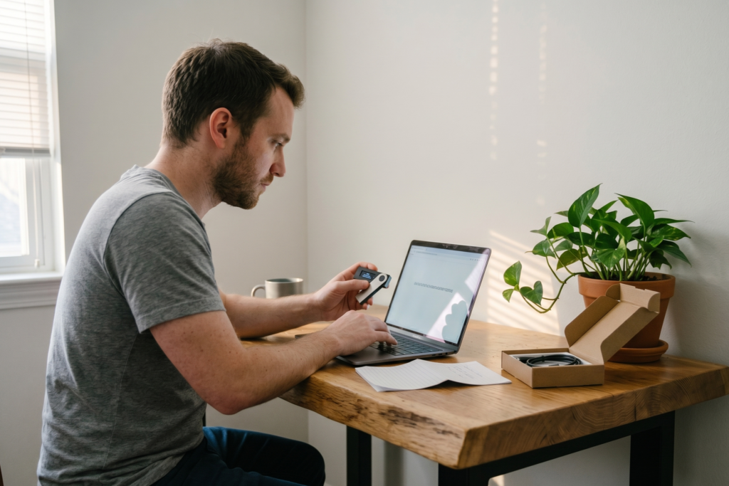 Man at home desk holding a hardware cold wallet while verifying a crypto address on his laptop screen.