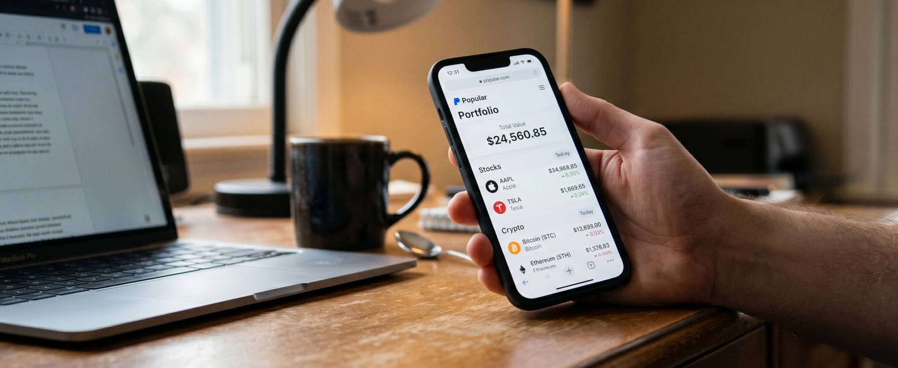 Hand holding smartphone showing Public.com portfolio app with Apple and Tesla stocks alongside Bitcoin and Ethereum crypto balances in sharp focus, with laptop and coffee mug softly blurred on wooden home office desk