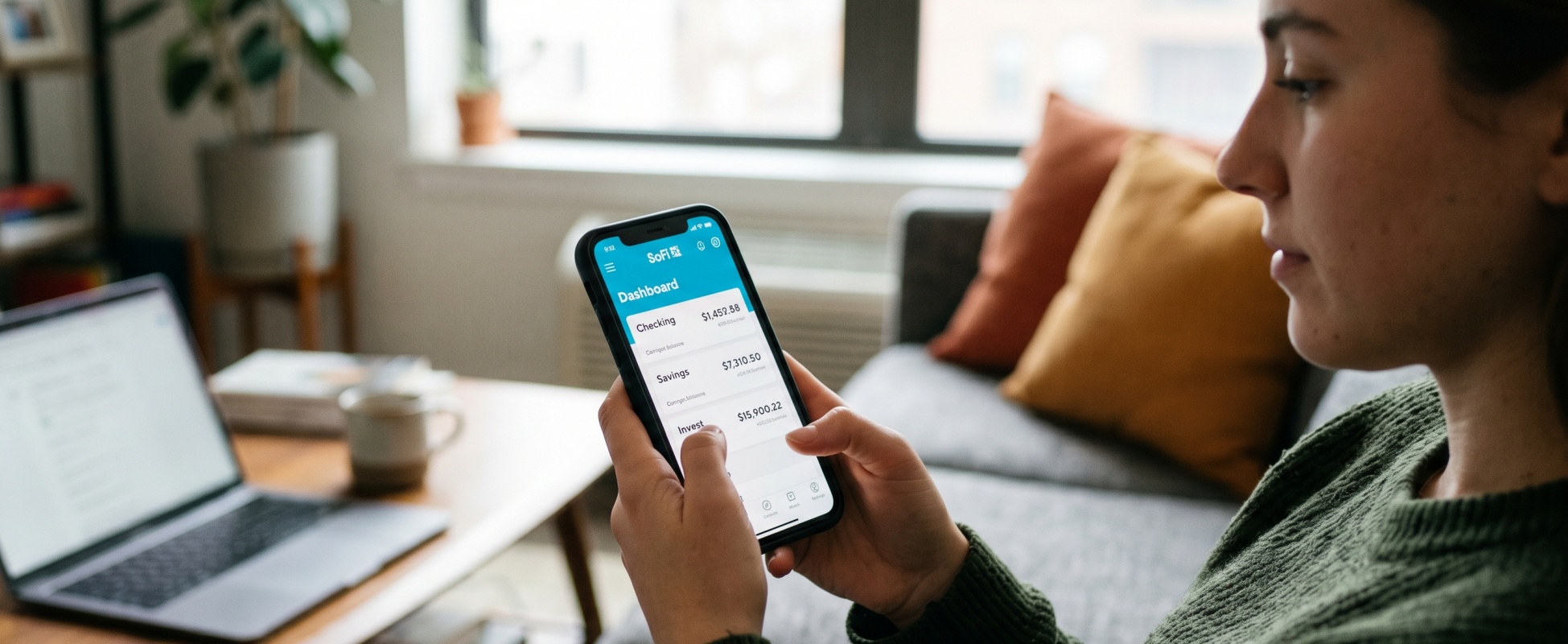 Young woman in green sweater holding smartphone showing SoFi banking app dashboard with checking, savings, and investing balances, with laptop and coffee mug on coffee table in modern apartment