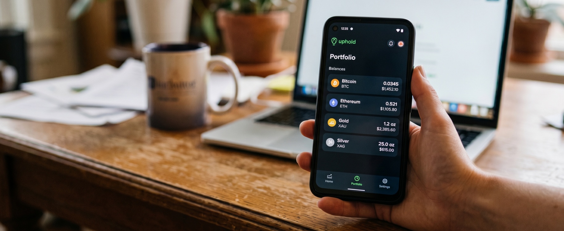 Hand holding smartphone showing Uphold portfolio app with Bitcoin, Ethereum, gold, and silver balances in sharp focus, with laptop and coffee mug softly blurred on wooden home office desk