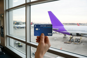 Hand holding a United Explorer credit card inside an airport terminal, with a purple-tailed airplane visible through the window.