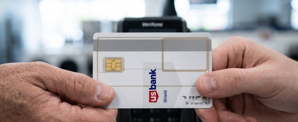Close-up of hands holding a white U.S. Bank Shield Visa credit card in front of a contactless payment terminal at an appliance store, with retail interior softly blurred in the background