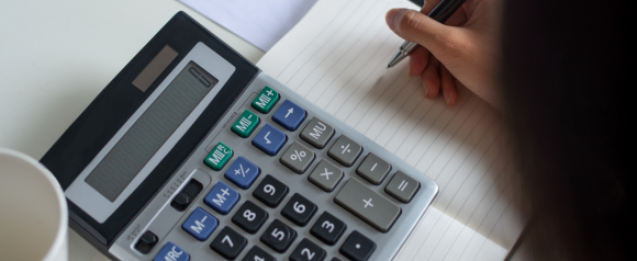 Close-up of a person writing in a lined notebook next to a large calculator, suggesting financial calculations or planning. The setup implies the detailed, numbers-driven work typical of tax law advocates.