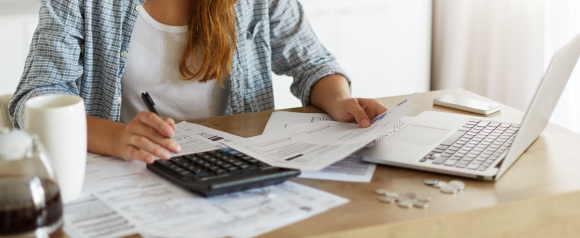 Woman sitting at a table using a calculator while reviewing bills and financial documents next to a laptop and a cup of coffee, symbolizing the process of managing debt and organizing paperwork for solutions like those offered by SoloSuit for debt relief.