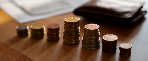 Stacks of assorted euro coins arranged in increasing height on a wooden table with a wallet and papers in the background, symbolizing budgeting and savings efforts often associated with programs like National Debt Relief.