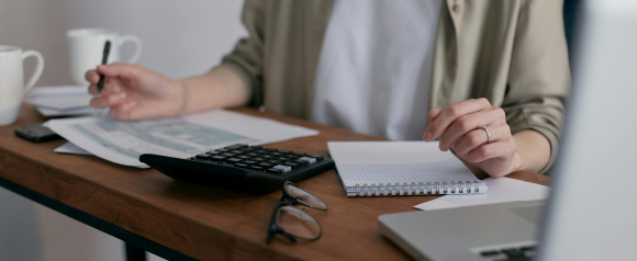 A woman sitting at a desk working with tax documents, a calculator, and a notebook, with a laptop and eyeglasses nearby. The scene reflects a personal finance or budgeting moment that aligns with Credit Saint debt relief services focused on financial organization and credit improvement.