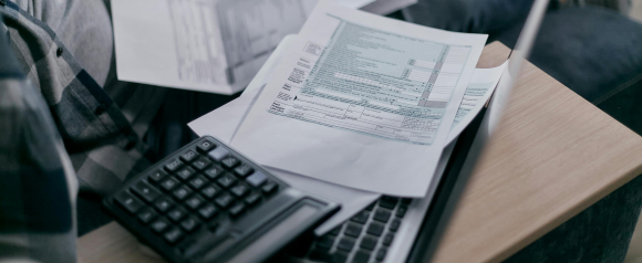 Close-up of tax forms and financial documents spread across a laptop and desk, with a calculator in the foreground. The image suggests someone working on finances or exploring debt relief options, possibly related to Americor services.