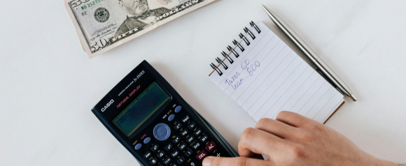 Hand using a Casio scientific calculator with a calculation displayed, next to a notepad reading “Taxes 60, Loan 800” and a stack of $50 bills, illustrating debt tracking and budgeting—key steps in programs like Freedom Debt Relief.