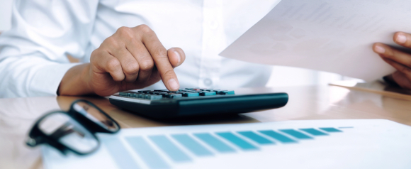 Close-up of a person in a white shirt using a calculator while holding a financial document, with a bar graph and eyeglasses on the desk. The image suggests financial planning or efforts to alleviate tax burdens through calculations and documentation.