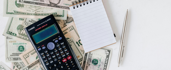 A scientific calculator, blank notepad, and silver pen placed on top of scattered U.S. dollar bills. The setup suggests financial planning or budgeting, relevant for services offered by tax relief experts.