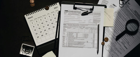 A close-up of various U.S. tax documents including Form 1040, W-7, and 4506-T scattered on a dark desk with a clipboard, black pen, coins, paperclips, magnifying glass, and a 2021 March calendar, suggesting a looming tax deadline. A sticky note labeled “tax expenses” hints at disorganized or complex tax prep, relevant to common 1099 tax problems.
