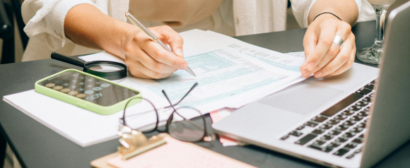 Close-up of a person filling out tax forms at a desk with a laptop, calculator, and magnifying glass, symbolizing attention to detail and paperwork involved in priority tax relief. Eyeglasses and a clipboard rest nearby, suggesting a focused work session.