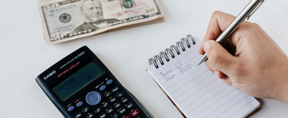 Close-up of a person writing “Taxes 60, Loan 800” in a notepad next to a scientific calculator displaying a math equation and a stack of $50 bills, reflecting tax and debt calculations similar to those handled by Optima Tax Relief.