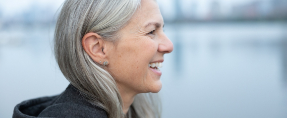 A smiling older woman with long gray hair and small earrings stands in profile by a calm waterfront. She wears a dark hoodie and leather jacket, with a soft cityscape blurred in the background. Suitable for content about hearing aids or active lifestyles for older adults, including Audicus-related topics.
